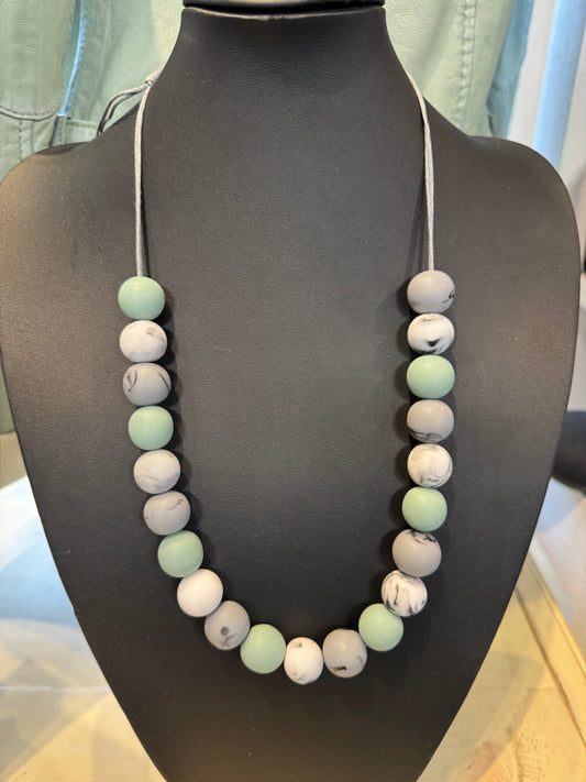 A versatile accessory from Suzie Blue, the Necklace Adjustable Marble - Blue/Grey features round beads in various shades of blue and gray displayed on a black mannequin bust. The marbled beads are strung on a thin, light-colored cord, with the background including a partial view of a garment.