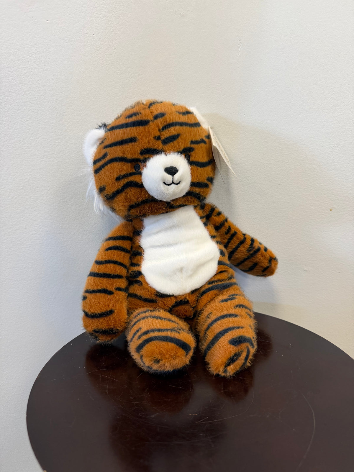 Certainly! Here’s the revised sentence using your product data: Gavin the Tiger – Minky by Fiesta, a plush toy tiger with black stripes, a white snout, and belly from Fiesta, sits on a dark wooden round table against a light gray wall.