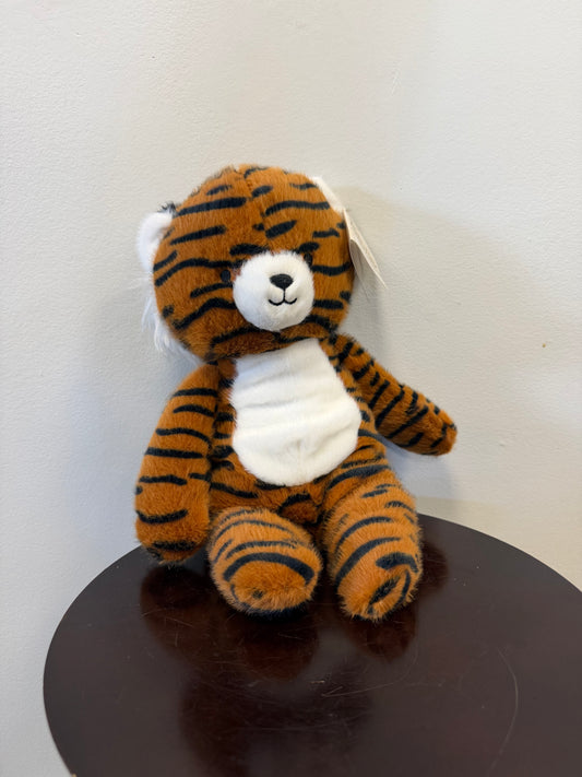 Certainly! Here’s the revised sentence using your product data: Gavin the Tiger – Minky by Fiesta, a plush toy tiger with black stripes, a white snout, and belly from Fiesta, sits on a dark wooden round table against a light gray wall.