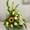 The "Warm Remembrance" funeral arrangement by Posies Flowers & Fashion features white roses, yellow blooms, a vibrant sunflower, and lush green foliage arranged in a white vase. A ribbon with "Grandpa" written on it adorns the bouquet. The arrangement is displayed on a gray surface against a light-colored wall next to a wooden box.