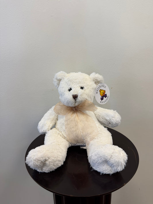 A Waterdale Classic Teddy Bear – White, crafted from recycled polyester and adorned with a beige ribbon around its neck, sits on a dark round table. The bear features a tag on its ear displaying the Waterdale bear logo and brand text. The background is a plain light-colored wall.