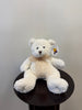 A Waterdale Classic Teddy Bear – White, crafted from recycled polyester and adorned with a beige ribbon around its neck, sits on a dark round table. The bear features a tag on its ear displaying the Waterdale bear logo and brand text. The background is a plain light-colored wall.