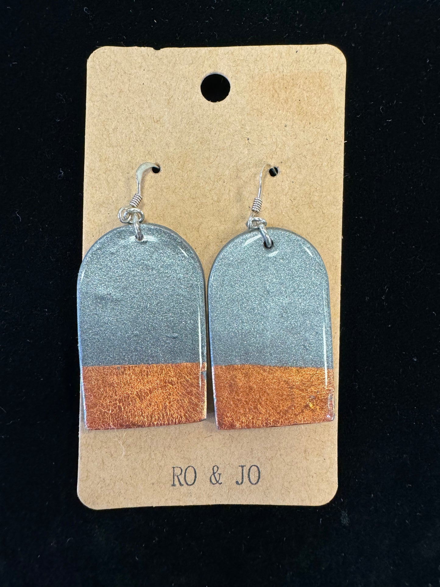 A pair of Ro&Jo Earrings 422 displayed on a brown rectangular card labeled "RO&JO." The earrings have a rounded rectangular shape with a silver top half and a bronze bottom half, attached to silver hooks. The background is black.