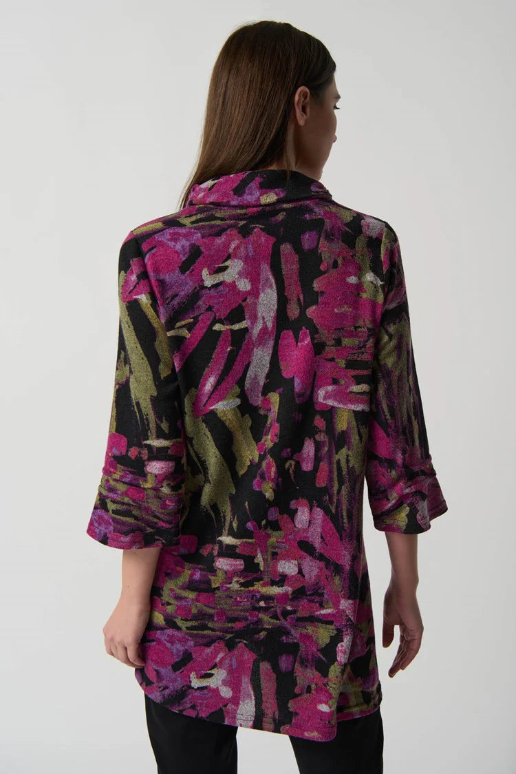 A woman with long brown hair is wearing the Joseph Ribkoff Black/Multi Printed Cowl Neck Tunic Style 233056, featuring an abstract pattern in black, pink, and green hues. She stands against a plain white background, with one hand resting in her pocket and a neutral expression on her face.