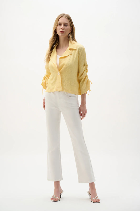 A woman with long, wavy blonde hair models the Joseph Ribkoff Woven Seersucker Boxy Blazer With Shirred Sleeves (262143) against a plain white background, paired with white cropped pants and white heels.