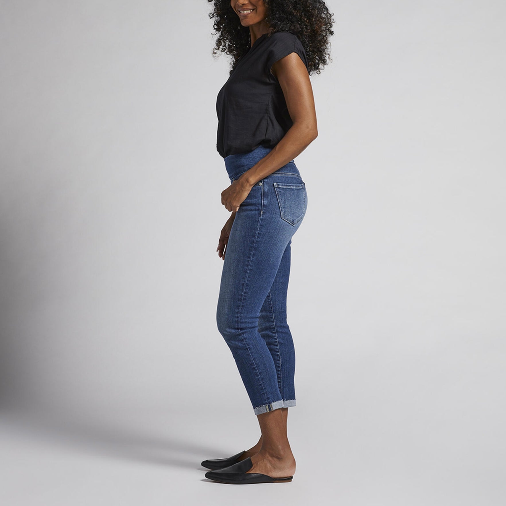 A person with curly hair is wearing a black short-sleeve blouse, Amelia Mid Rise Slim Ankle Pull-On Jeans by JAG, and black loafers. They are standing against a plain light grey background, smiling, with their hands relaxed by their sides.