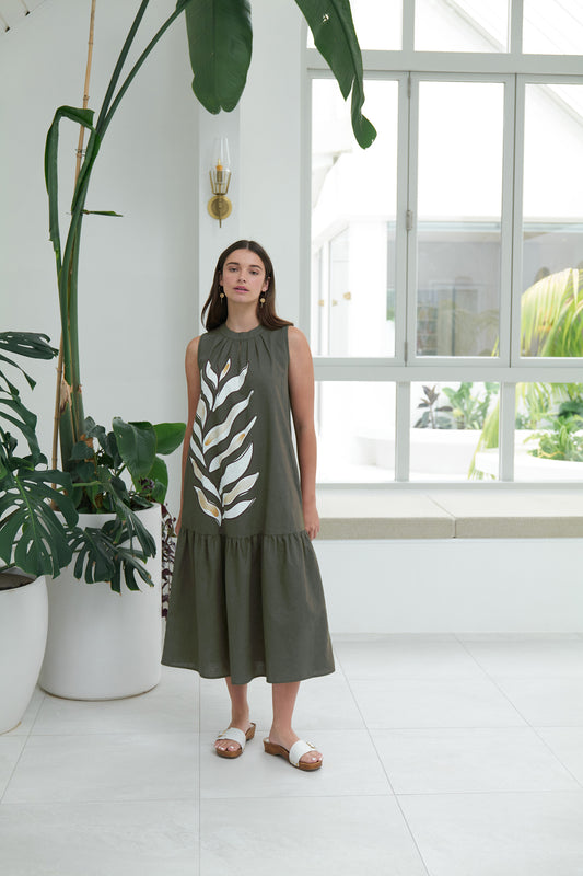 A woman wears the FOIL To The Max Dress FO8170 Palm Life, a sleeveless tropical leaf-print dress, with white sandals. She stands indoors among tall potted plants, surrounded by white walls and large windows letting in natural light.