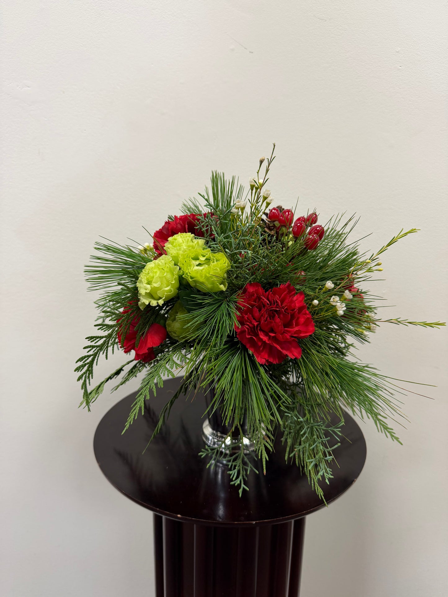 The "Little Cup of Christmas" by Posies Flowers & Fashion features red and green blooms, pine needles, and red berries in a glass vase, perfect for festive decor against a light-colored wall.