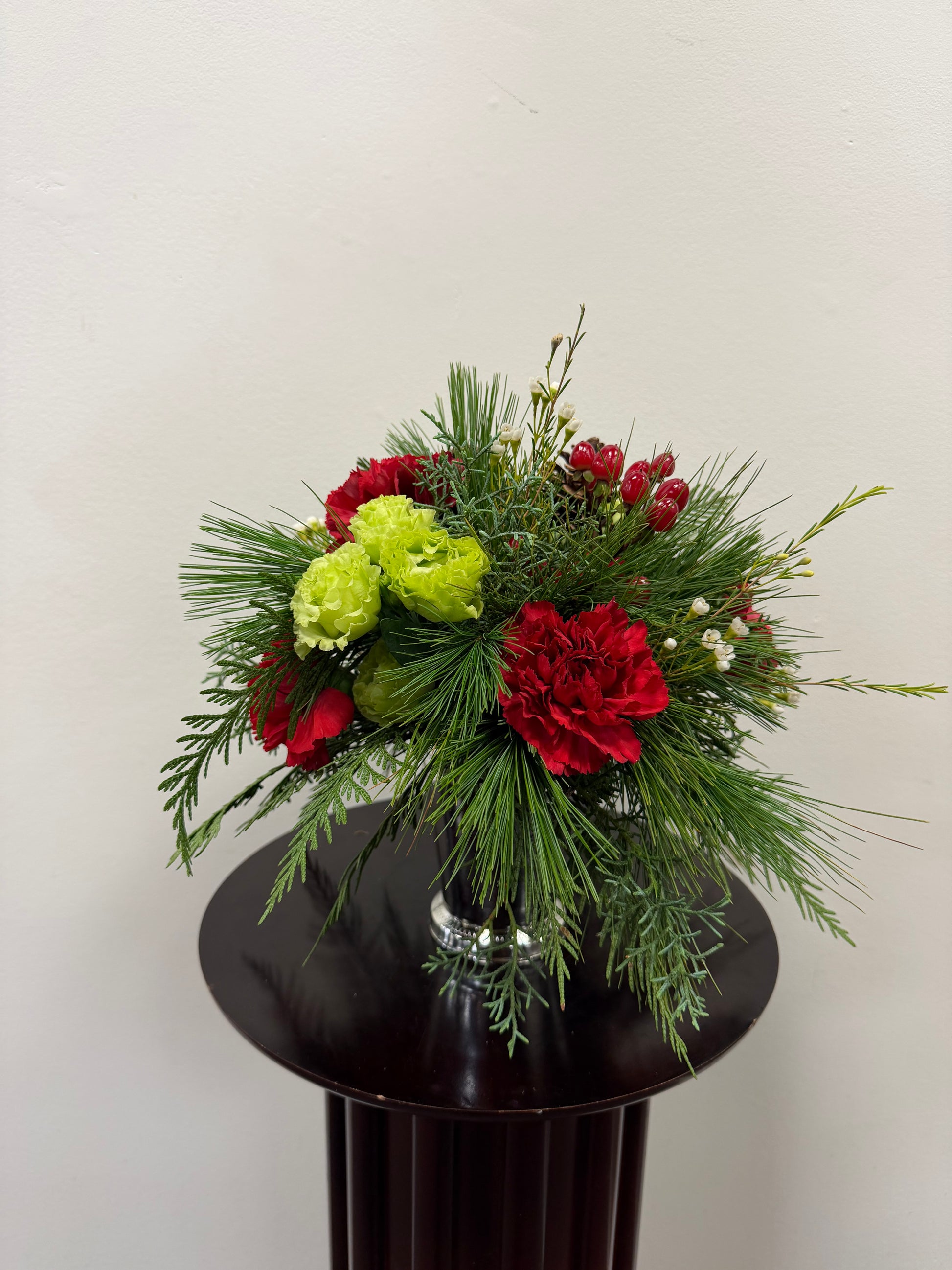 The "Little Cup of Christmas" by Posies Flowers & Fashion features red and green blooms, pine needles, and red berries in a glass vase, perfect for festive decor against a light-colored wall.