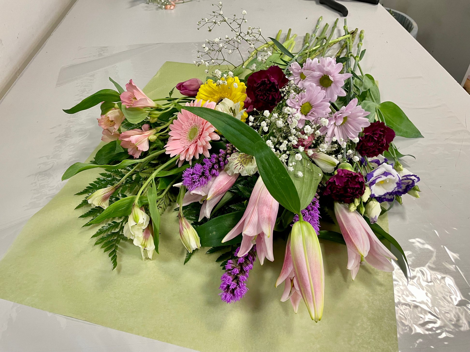 A stunning bouquet called Designer’s Choice Freshcuts from Posies Flowers & Fashion is resting on a white surface. The arrangement features vibrant orange and pink roses, cheerful yellow chrysanthemums, elegant white flowers, and assorted green foliage. It is partially wrapped in brown paper and elegantly tied with a burgundy ribbon.