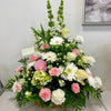 The "Peaceful Embrace" by Posies Flowers & Fashion is a beautifully arranged floral bouquet featuring a serene mix of delicate pink and white flowers, including roses, gerberas, carnations, and other blossoms. This arrangement is surrounded by green foliage and exquisitely displayed in a wooden container against a light-colored wall.