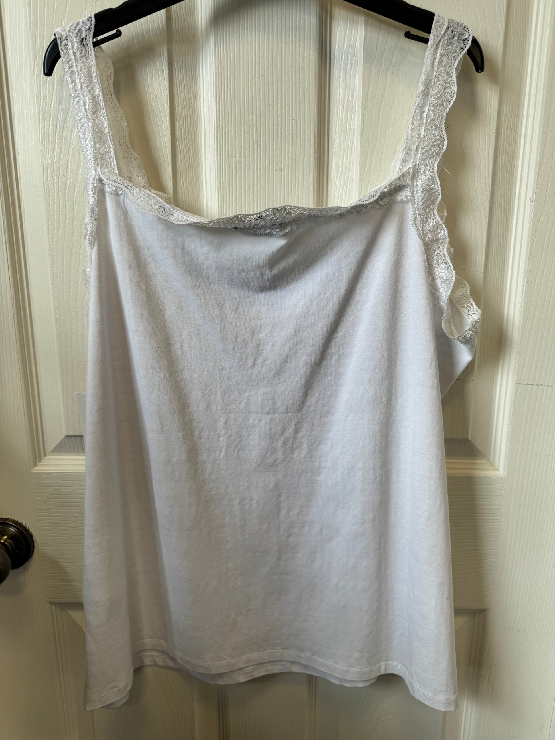 A classic "Tribal Cami White - 1516O-403-0001" with wide lace-trimmed straps hangs on a black hanger against a white door. Made of 95% cotton, it features a price tag and product information tag attached to the right side.