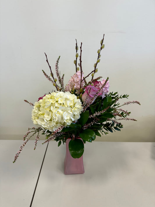 A Designer’s Choice Vase from Posies Flowers & Fashion, filled with white lilies and green foliage, graces a marble table in an elegantly modern room. Soft lighting and contemporary furniture in the background create a warm and inviting atmosphere, showcasing the artistry of floral designers, subject to availability.