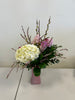 A Designer’s Choice Vase from Posies Flowers & Fashion, filled with white lilies and green foliage, graces a marble table in an elegantly modern room. Soft lighting and contemporary furniture in the background create a warm and inviting atmosphere, showcasing the artistry of floral designers, subject to availability.