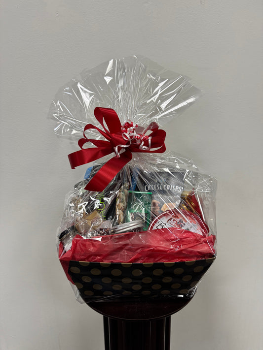 A Gift Basket from Posies Flowers & Fashion is elegantly wrapped in clear plastic adorned with a red bow. Inside, you'll find an assortment of snacks, featuring cheese crisps and mixed nuts, all beautifully arranged in a black box adorned with gold polka dots. The basket is displayed against a plain white background.