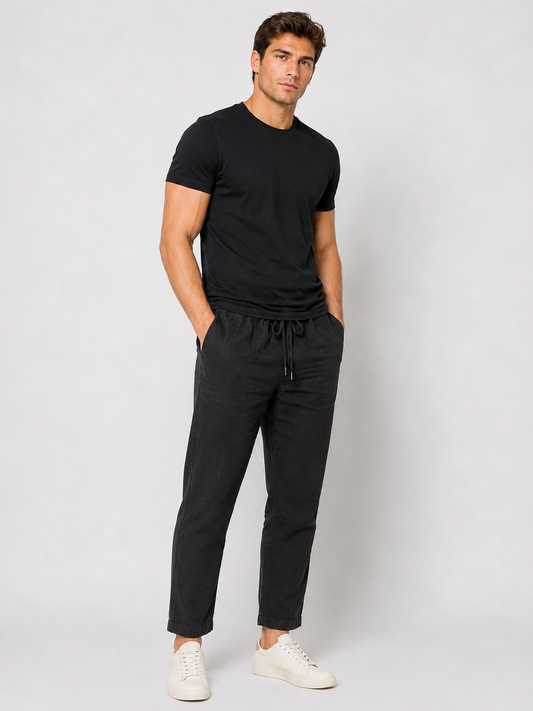 A man stands against a plain light background wearing a black t-shirt, Paul Miranda Linen Pant Nero PE680 with a drawstring, and white sneakers. His hands are in his pockets and he has a relaxed expression.