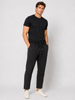 A man stands against a plain light background wearing a black t-shirt, Paul Miranda Linen Pant Nero PE680 with a drawstring, and white sneakers. His hands are in his pockets and he has a relaxed expression.