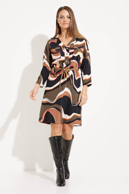 A woman stands against a white background, wearing the Abstract Print Shirt Dress Style 233224 by Joseph Ribkoff, which features wavy, multicolored patterns in beige, brown, and white. The long-sleeve dress has a belted waist and buttons down the top half. She is also wearing black knee-high boots.