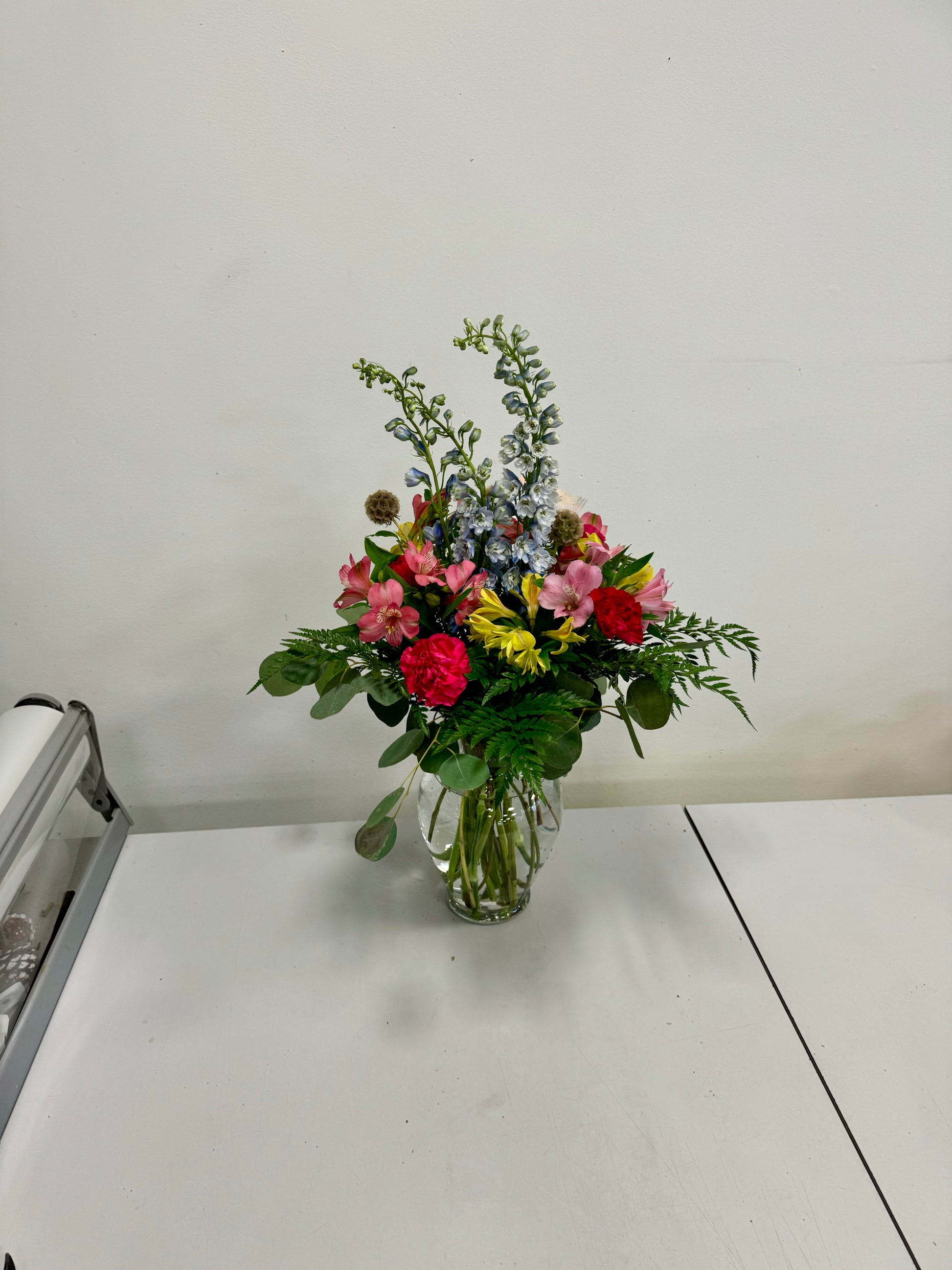 The "Spring Beauty" floral arrangement by Posies Flowers & Fashion graces a white table in a clear vase. This vibrant bouquet, positioned prominently in the image, features an assortment of pink, red, yellow, and blue blossoms with green foliage. Although flower availability might necessitate substitutions, the display remains colorful and fresh against the plain white background.