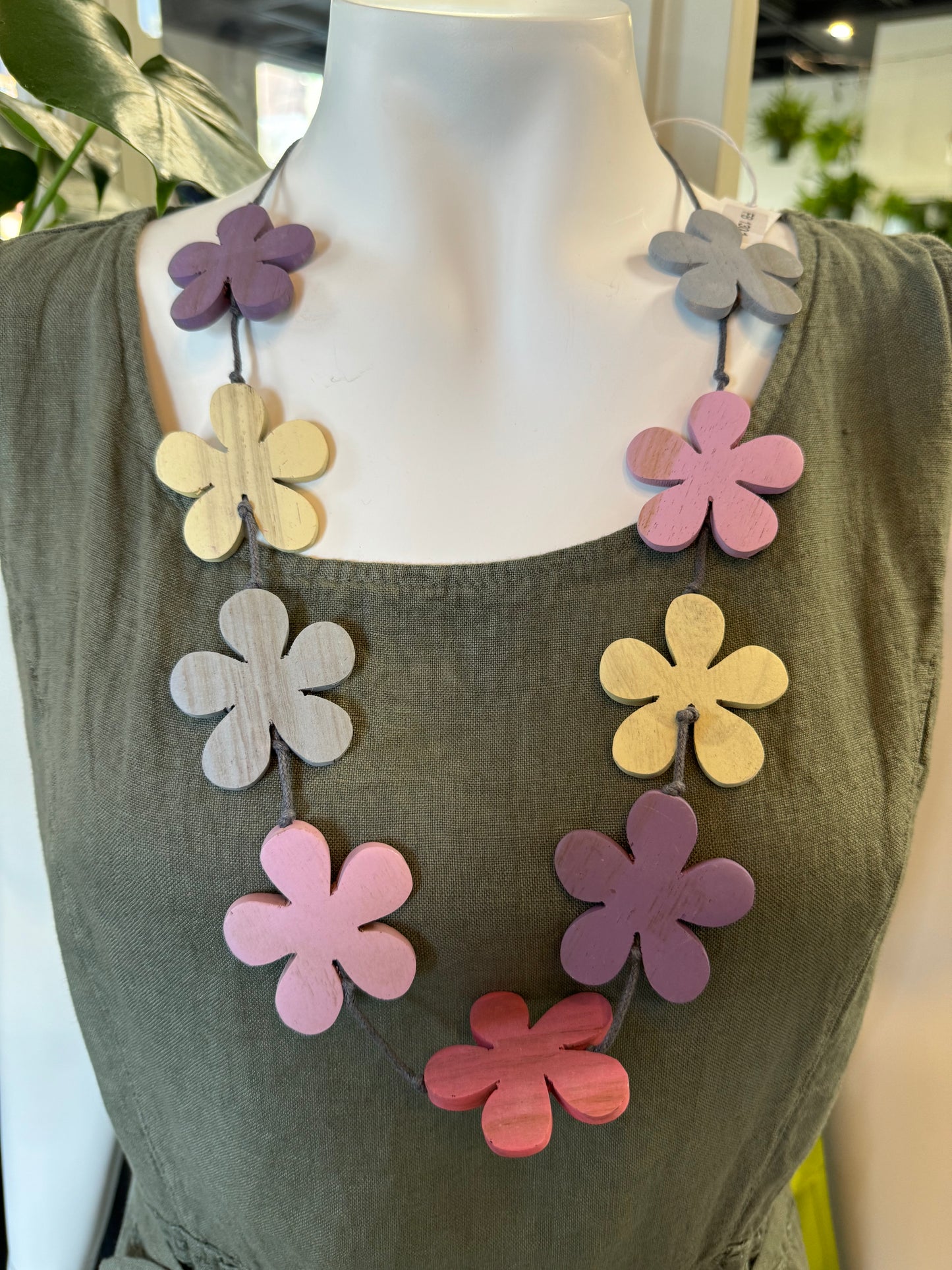 A Suzie Blue Purple Shades Flower Necklace rests on a textured carpeted surface. This handcrafted piece showcases an arrangement of elegant, wooden, flower-shaped beads in pastel colors, including pink, purple, yellow, and gray, strung on a cord with black bead details. A small tag is attached to the necklace.