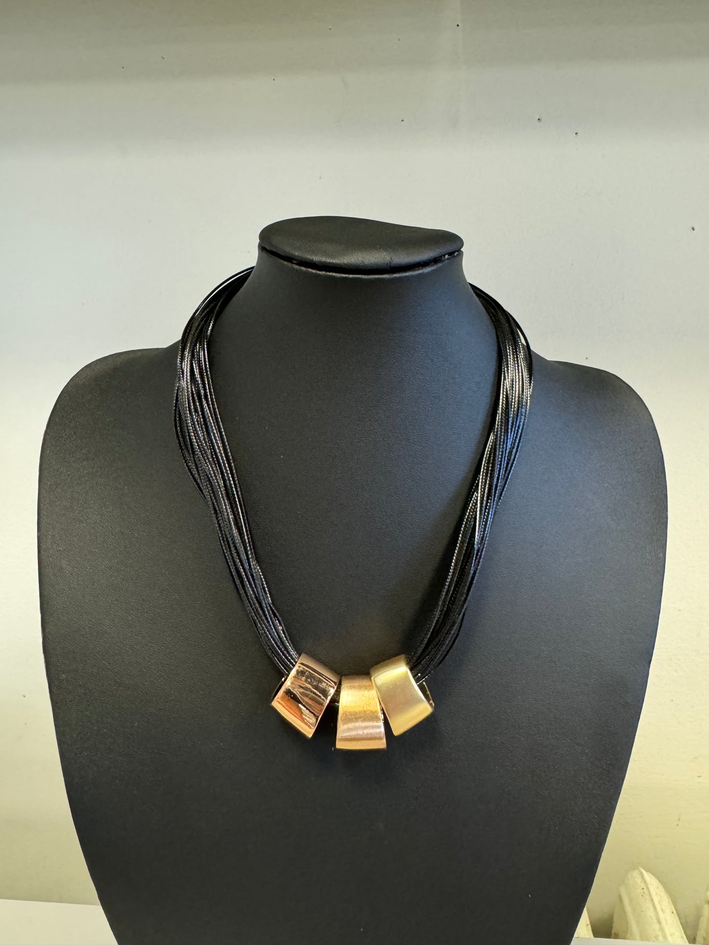 The Roxanne 28 Necklace by Elise Accessories is a stunning fashion accessory showcasing a black multi-strand design with three elegant gold square-shaped pendants, beautifully presented on a black bust.