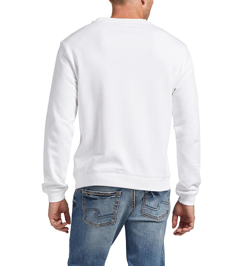 A man is modeling the Silver Jeans Co. Mens Crewneck Sweatshirt in white, featuring subtle embroidered text "49.8951°N 97.1384°W" on the front. He pairs it with light blue jeans, has short hair, and is looking to his left against a plain white background.