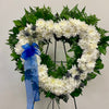 The "Eternal Serenity" wreath by Posies Flowers & Fashion is heart-shaped and crafted from white carnations and green leaves, elegantly displayed on a black metal easel. This serene arrangement is adorned with a prominent blue ribbon bow on the left side, accented with scattered blue thistle flowers among the white blooms.