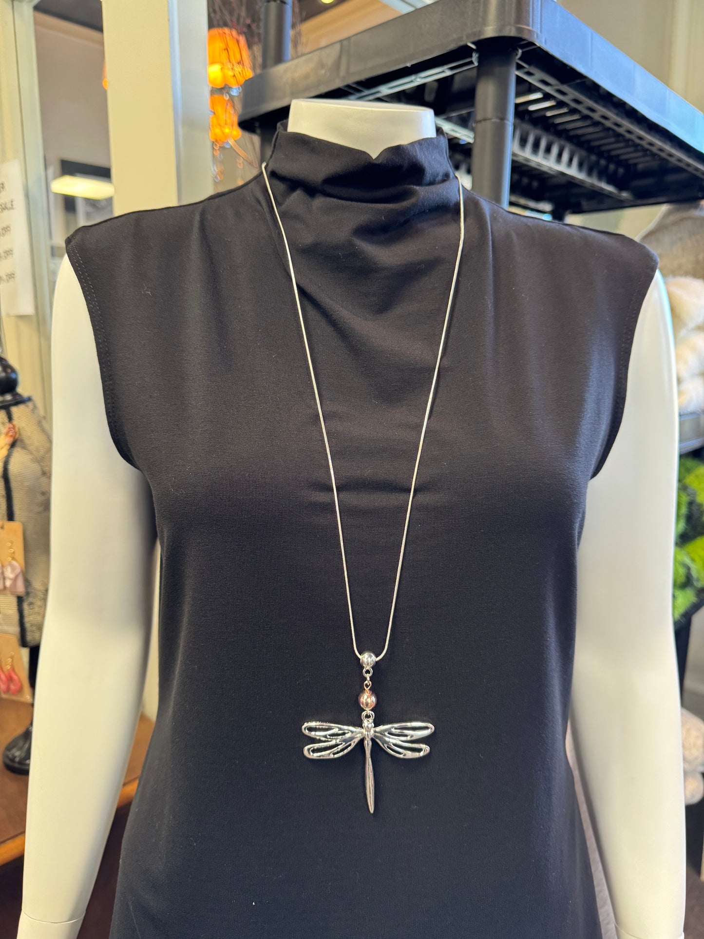 A mannequin displays an elegant, sleeveless black turtleneck dress, beautifully accentuated by the long Necklace 10 from Elise Accessories, which features a striking silver dragonfly pendant. The stylish ensemble is showcased in a chic retail display area.