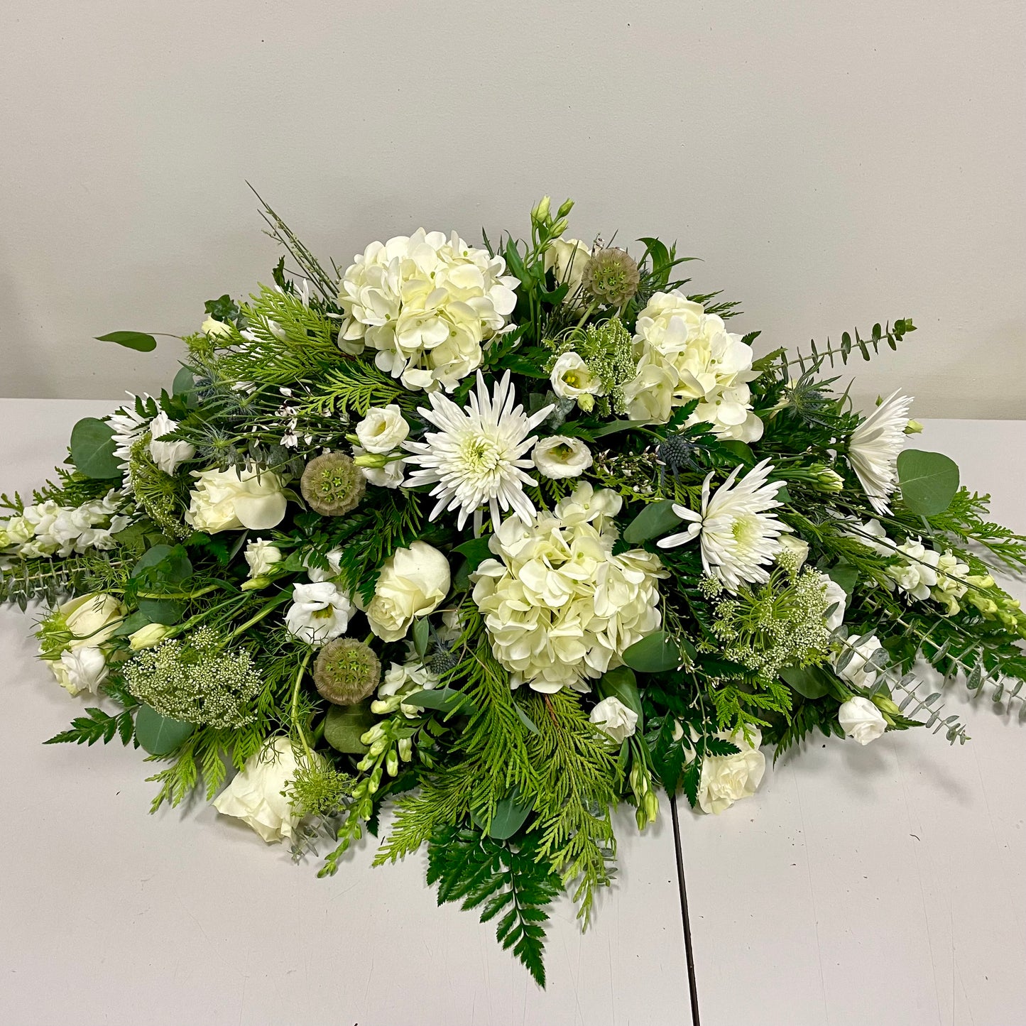 The *Expression in White* by Posies Flowers & Fashion is a stunning floral arrangement featuring white roses, daisies, and other captivating white blooms, accented with an array of green foliage. These flowers are meticulously arranged on a plain, light-colored surface against a neutral background—making it an ideal choice for a serene open casket display or an elegant casket spray.