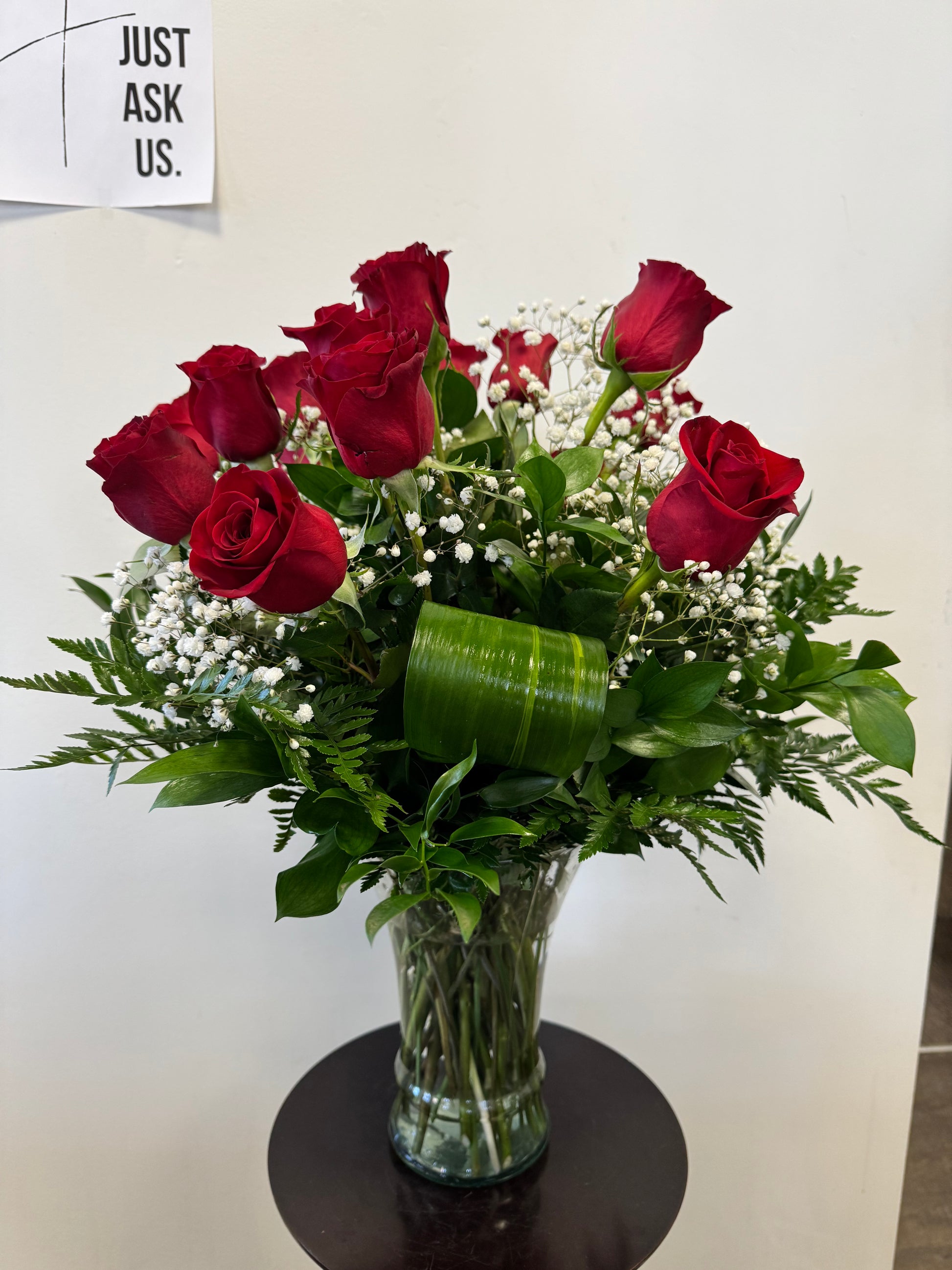 The Roses Roses Vase by Posies Flowers & Fashion features red roses, baby's breath, and greenery in a clear vase on a round dark table. Flower availability may vary; similar substitutions may occur. A "JUST ASK US" sign is on the wall.