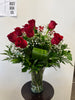The Roses Roses Vase by Posies Flowers & Fashion features red roses, baby's breath, and greenery in a clear vase on a round dark table. Flower availability may vary; similar substitutions may occur. A "JUST ASK US" sign is on the wall.