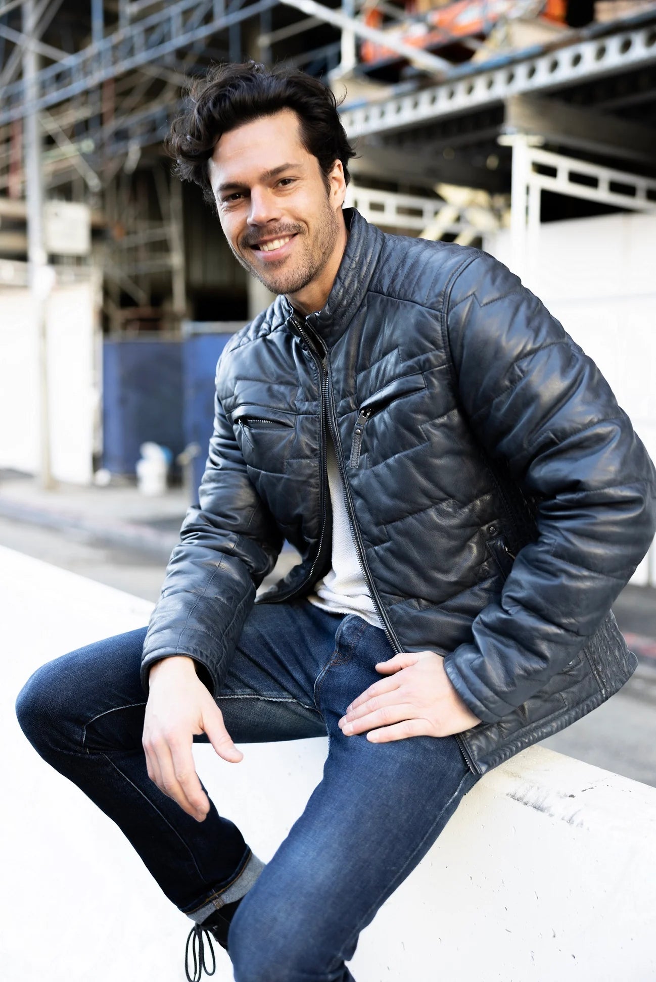 A man with short, dark hair, wearing a stylish navy Aplin CF Leather Jacket by MAURITIUS LEATHER, along with a white shirt and blue jeans, is smiling and sitting on a concrete ledge outdoors. There is a metal scaffolding structure in the background.