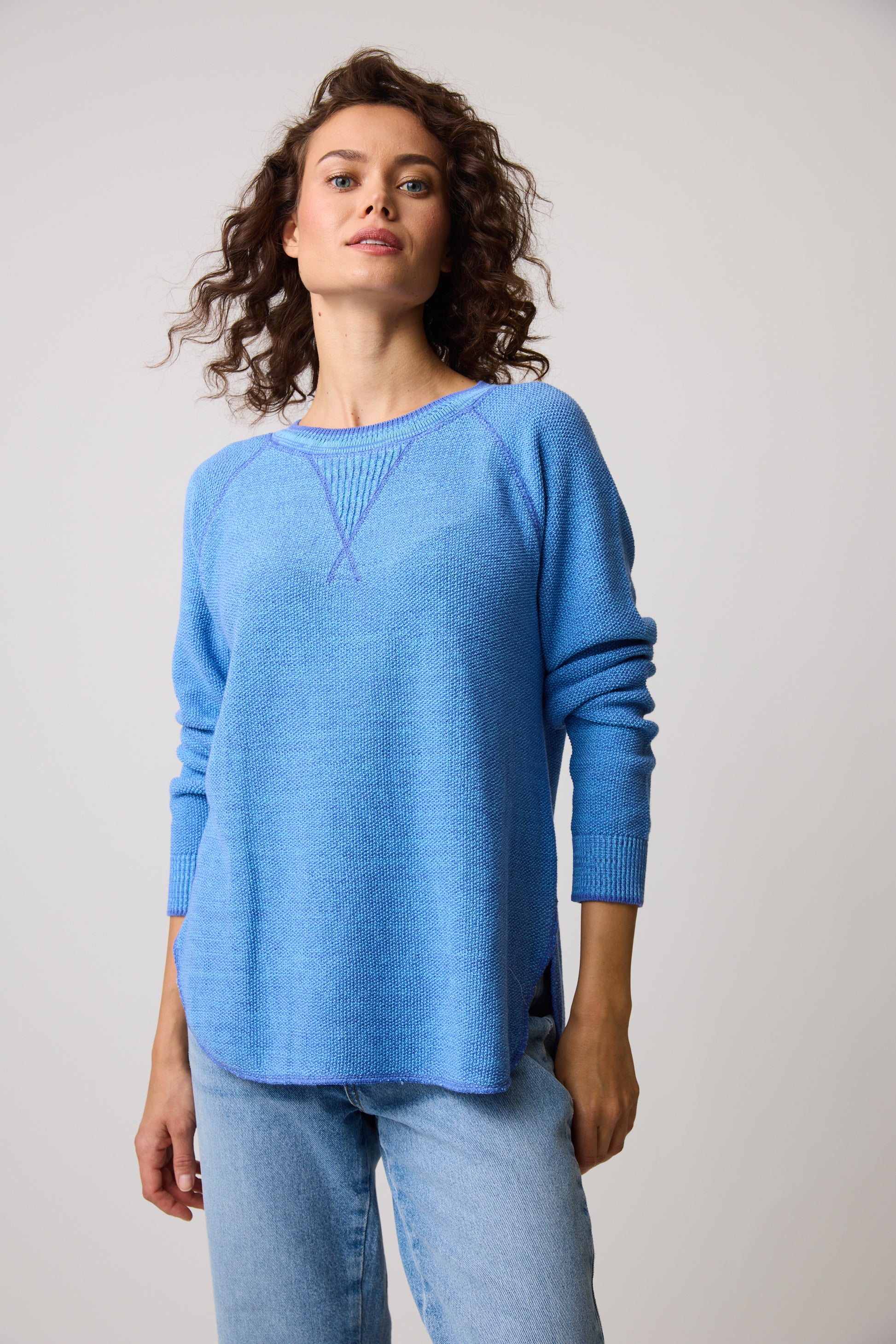 A person with curly hair wears the Skyler Sweatshirt 80049 from Parkhurst Cotton Country in light blue, made of 100% cotton, paired with jeans. They stand against a plain backdrop, gazing upward calmly.