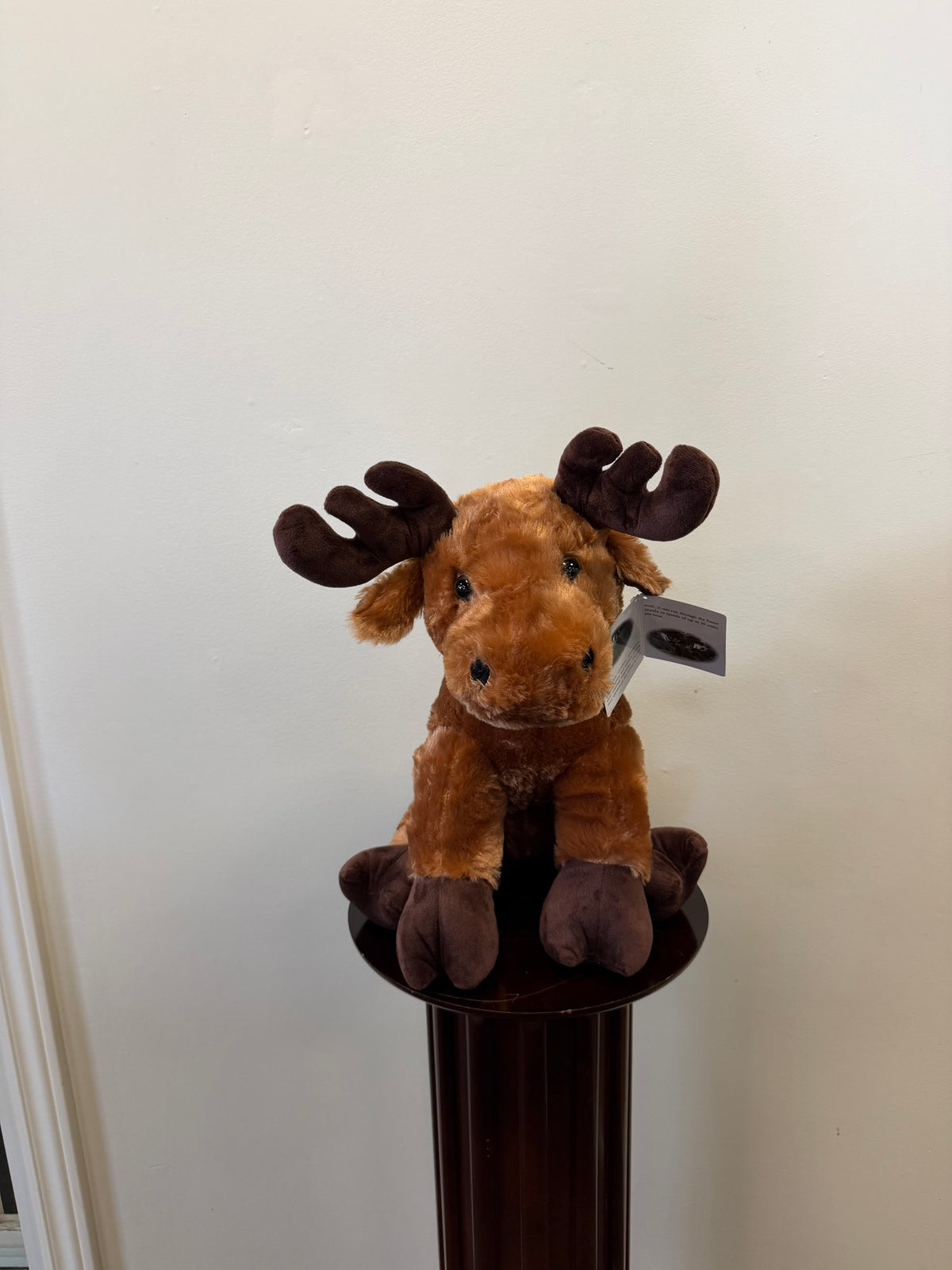 The Fiesta Plush Moose - 15 inch features dark brown antlers and sits on a round, dark wooden pedestal against a plain white wall.