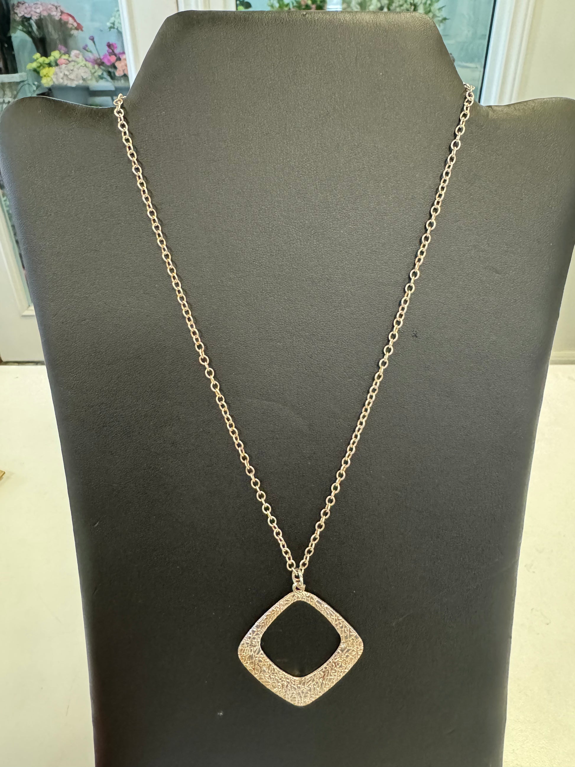 A stunning piece from Suzie Blue, the Rose Gold Coloured Necklace with a large diamond-shaped pendant is displayed on a black jewelry stand, exuding luxury against a blurred indoor setting backdrop.