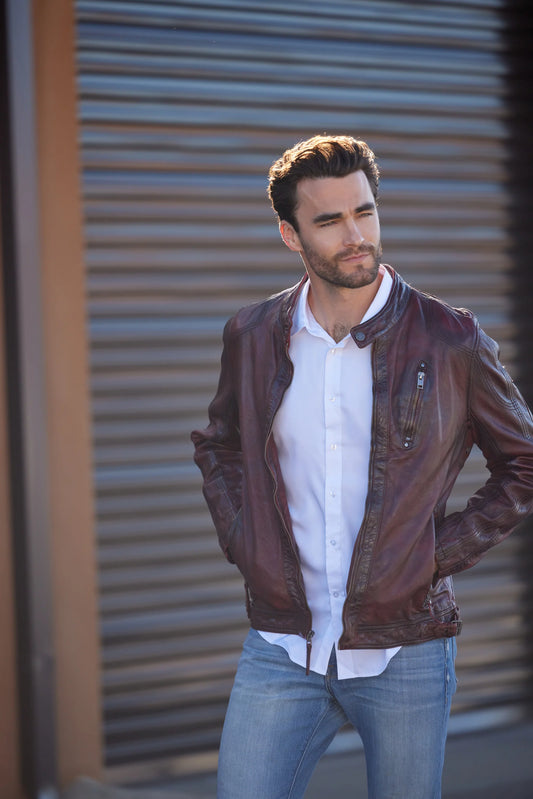 A man with dark hair and a beard stands in front of a corrugated metal background, wearing a white dress shirt and a premium quality MAURITIUS LEATHER Rakva RF Leather Jacket in wine, with his hands in his jacket pockets.