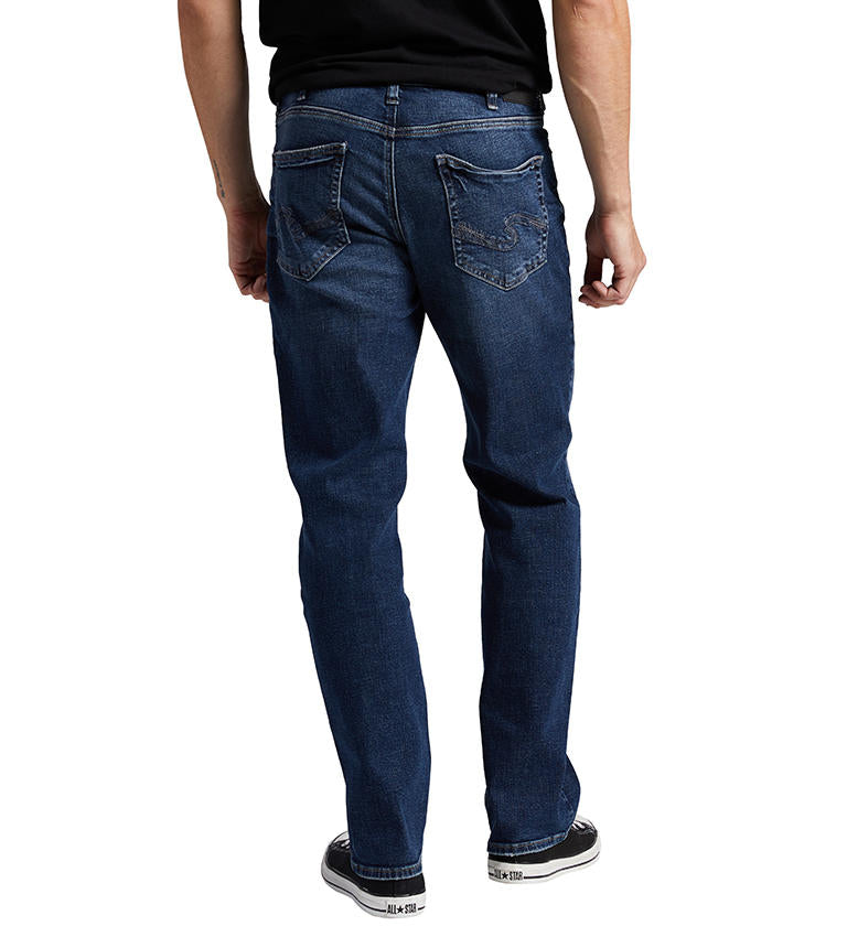 A person is standing, wearing Silver Jeans Co.'s Grayson Classic Fit Straight Leg Jeans in a dark indigo wash with classic five-pocket styling. The slightly faded straight-leg jeans are paired with a black shirt and black low-top sneakers with white soles. The background is white.