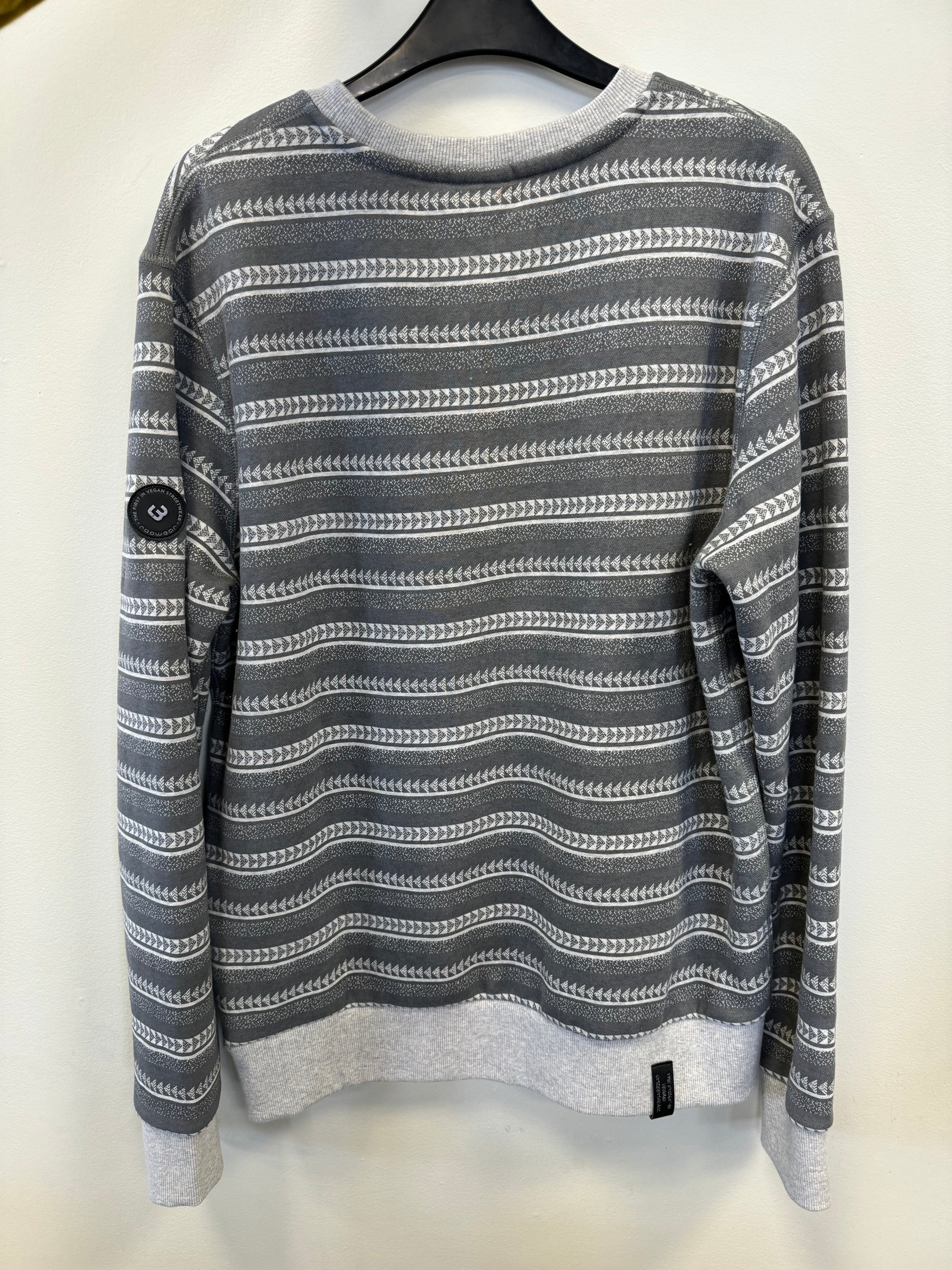 A Ragwear Sweatshirt Pullover - INDDIE PRINT by RAGWEAR, featuring gray and white stripes with a pattern of horizontal wavy lines and white geometric shapes, is hanging on a black hanger against a white wall. The vegan sweatshirt also has gray cuffs and hem, and a small black tag with writing is sewn on the front.