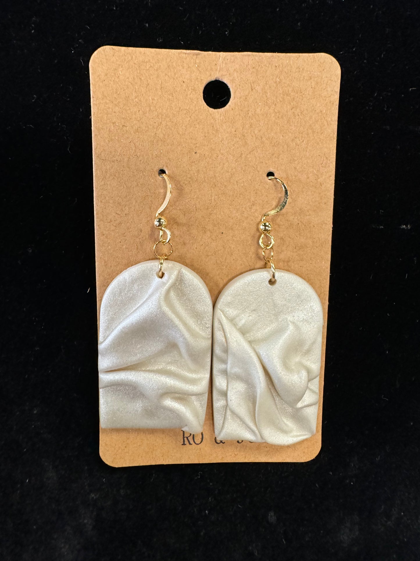 Introducing the Ro&Jo Earrings 443 from RO&JO: a pair of cream-colored, handmade earrings with a distinctive wrinkled texture. Each arched earring elegantly hangs from a small, gold-colored hook. These unique jewelry pieces come beautifully displayed on a rectangular brown card with a hole at the top for hanging.