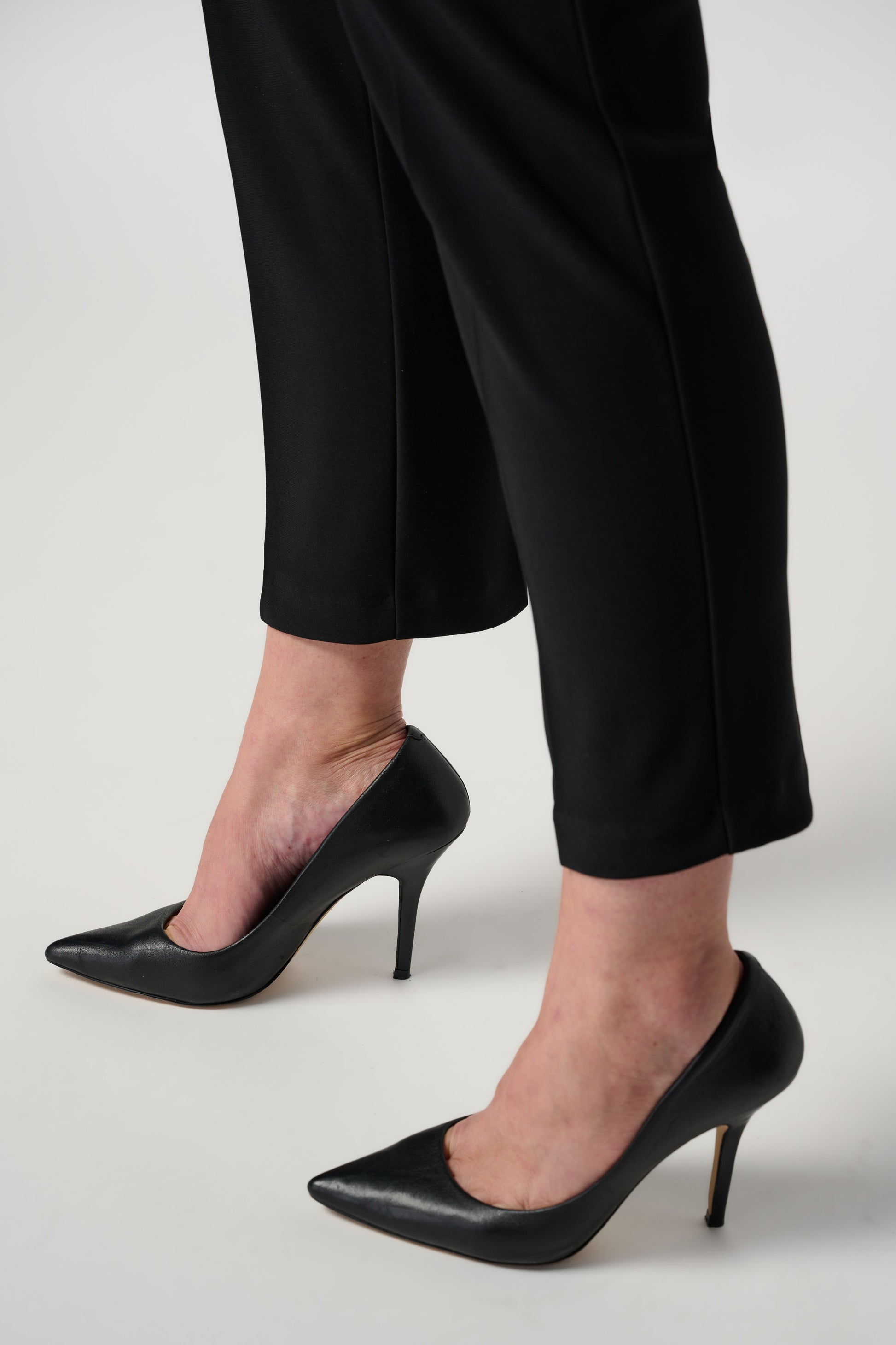 A person is donning Joseph Ribkoff's Classic Cropped Pant paired with black high-heeled shoes. The top portion of their outfit is a white shirt, and their arms rest by their sides. The background is plain and white.