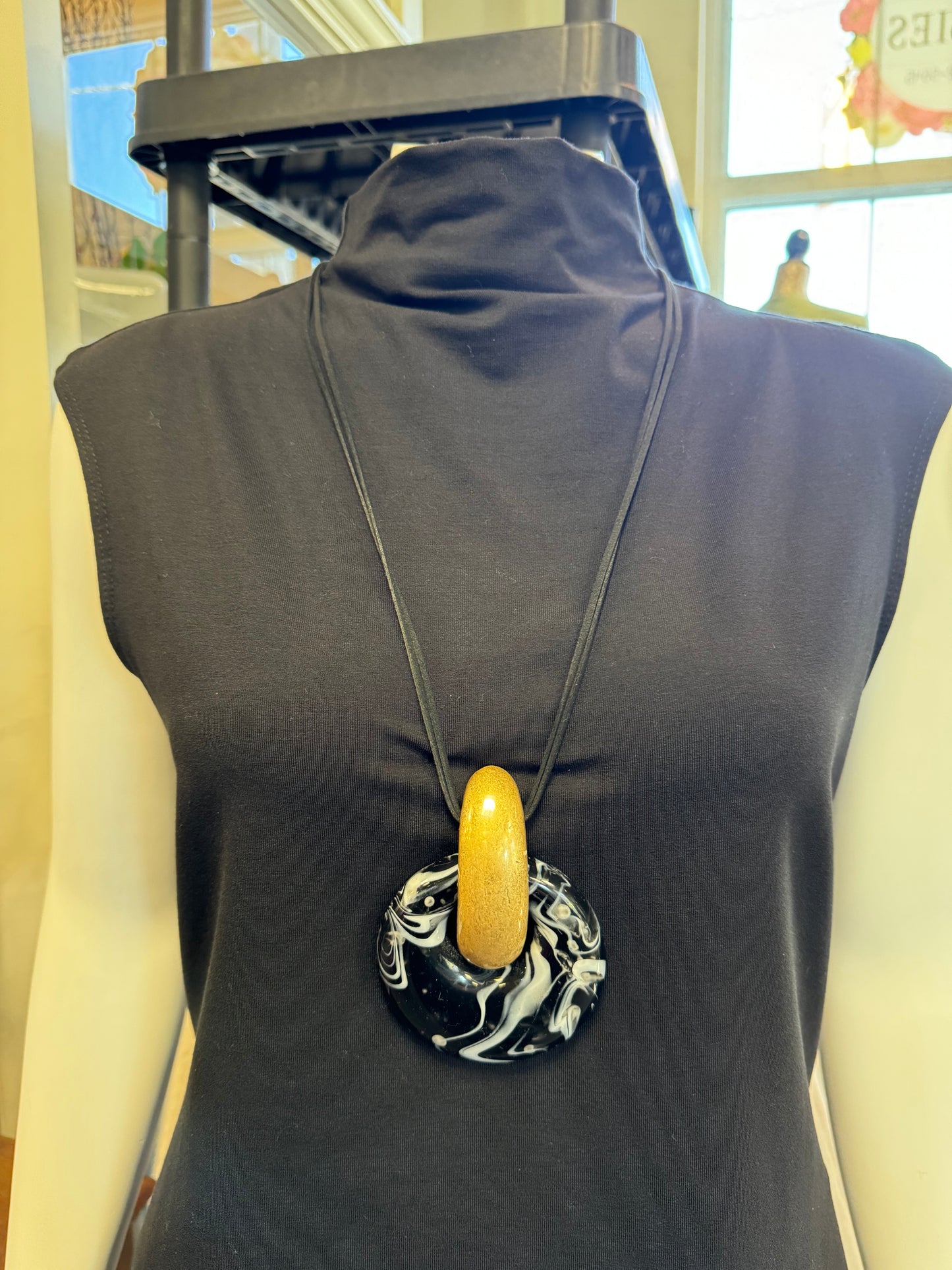 A mannequin dressed in a black sleeveless top elegantly showcases the fashion-forward Necklace from Elise Accessories. This piece features a large pendant with a shiny, marble-like design in black and white, crowned by a gold, crescent-shaped accent.