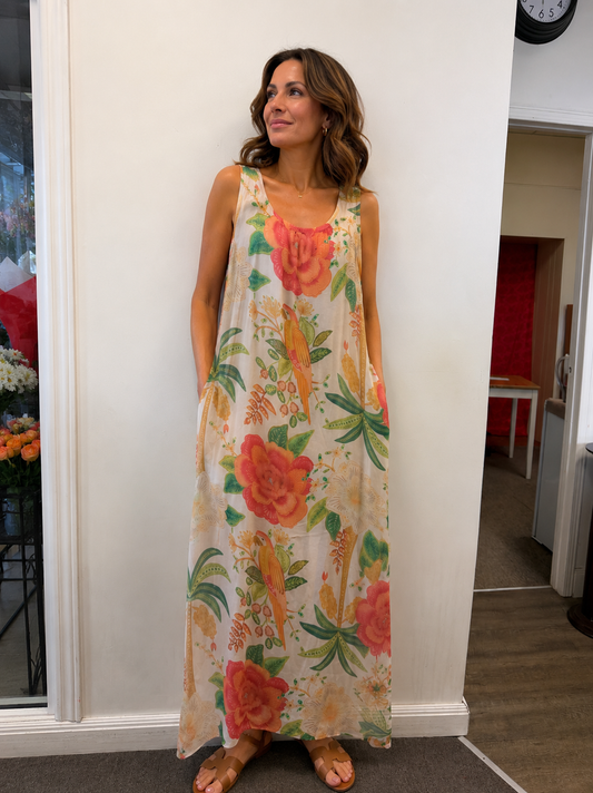 A woman stands indoors wearing the ETERNELLE Silk Blend Dress Sleeveless Sand (6-SS26174-9) with a bold floral pattern and brown sandals, looking to the side with her hands in her dress pockets.