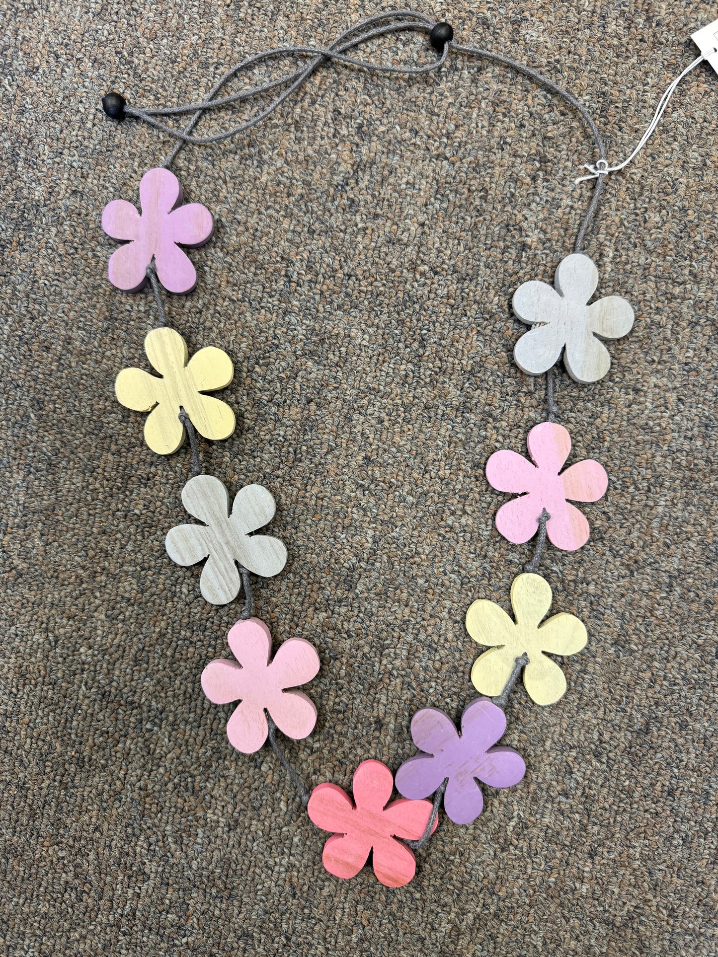 A Suzie Blue Purple Shades Flower Necklace rests on a textured carpeted surface. This handcrafted piece showcases an arrangement of elegant, wooden, flower-shaped beads in pastel colors, including pink, purple, yellow, and gray, strung on a cord with black bead details. A small tag is attached to the necklace.