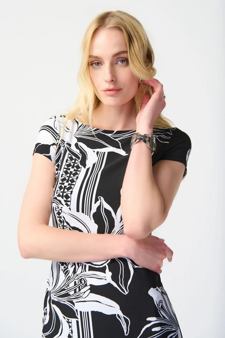 A woman with long blonde hair stands against a plain background, wearing the Joseph Ribkoff Floral Print Silky Knit Sheath Dress 241284. This short-sleeve black dress features a chic silhouette with large white flowers and geometric accents along the border. She has a neutral expression and one hand placed on her hip.