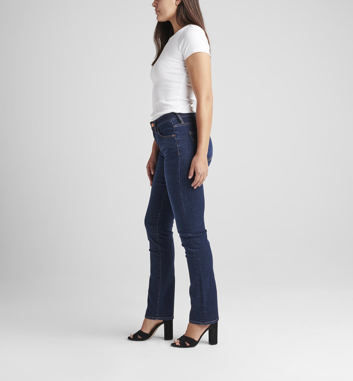 A person with long dark hair stands against a plain light gray background. They are wearing a short-sleeved white t-shirt, Ruby Mid Rise Straight Leg Jeans in dark blue power stretch denim by JAG, and black open-toe heels. The person holds one hand in their jeans pocket and stands with a slight, relaxed lean.