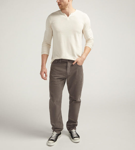 A person stands against a plain background, wearing a long-sleeve off-white henley shirt, corduroy pants in graphite from Silver Jeans Co. (Style M63915CCC649), and black low-top sneakers with white laces and soles. The person's head is turned slightly to the left, with their left arm hanging by their side and their right hand in their pocket.