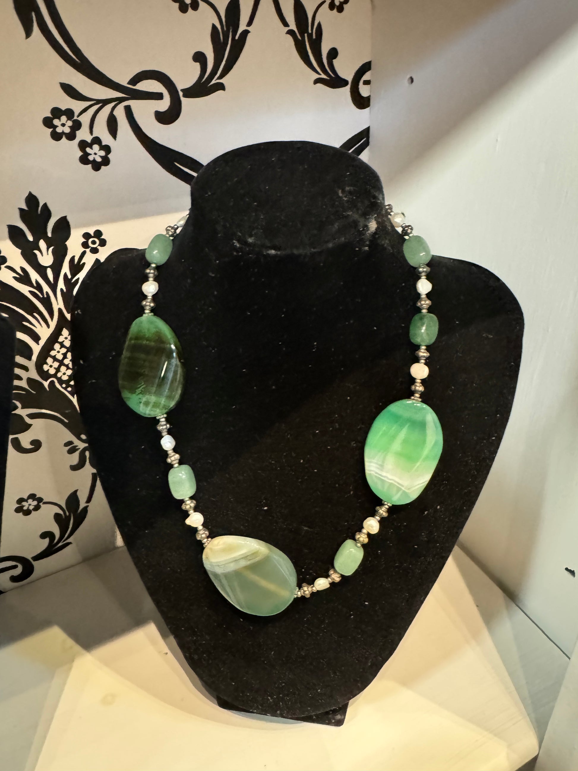 A black velvet jewelry display from Posies Flowers & Fashion Inc. showcases an elegant Turquoise, White, and Grey Coloured Necklace with Earrings, featuring large green and white striped oval stones, smaller green beads, and small white pearls. The background has a decorative black and white floral pattern on the left side, highlighting the beauty of this coloured necklace.