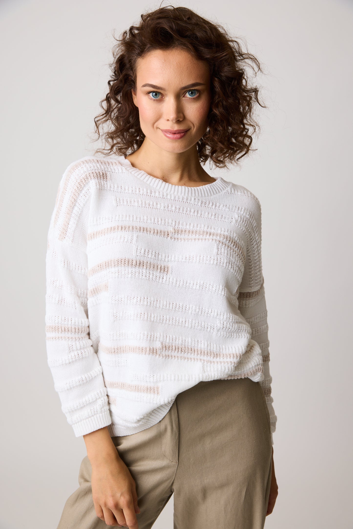 A person with curly hair is smiling at the camera, wearing a white Abigail Stripe Alabaster 87328 sweater from Parkhurst Cotton Country and beige pants against a plain, light-colored background.