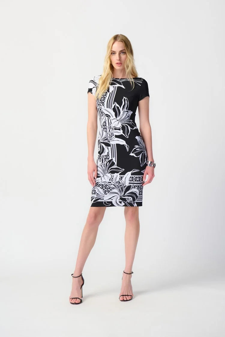 A woman with long blonde hair stands against a plain background, wearing the Joseph Ribkoff Floral Print Silky Knit Sheath Dress 241284. This short-sleeve black dress features a chic silhouette with large white flowers and geometric accents along the border. She has a neutral expression and one hand placed on her hip.