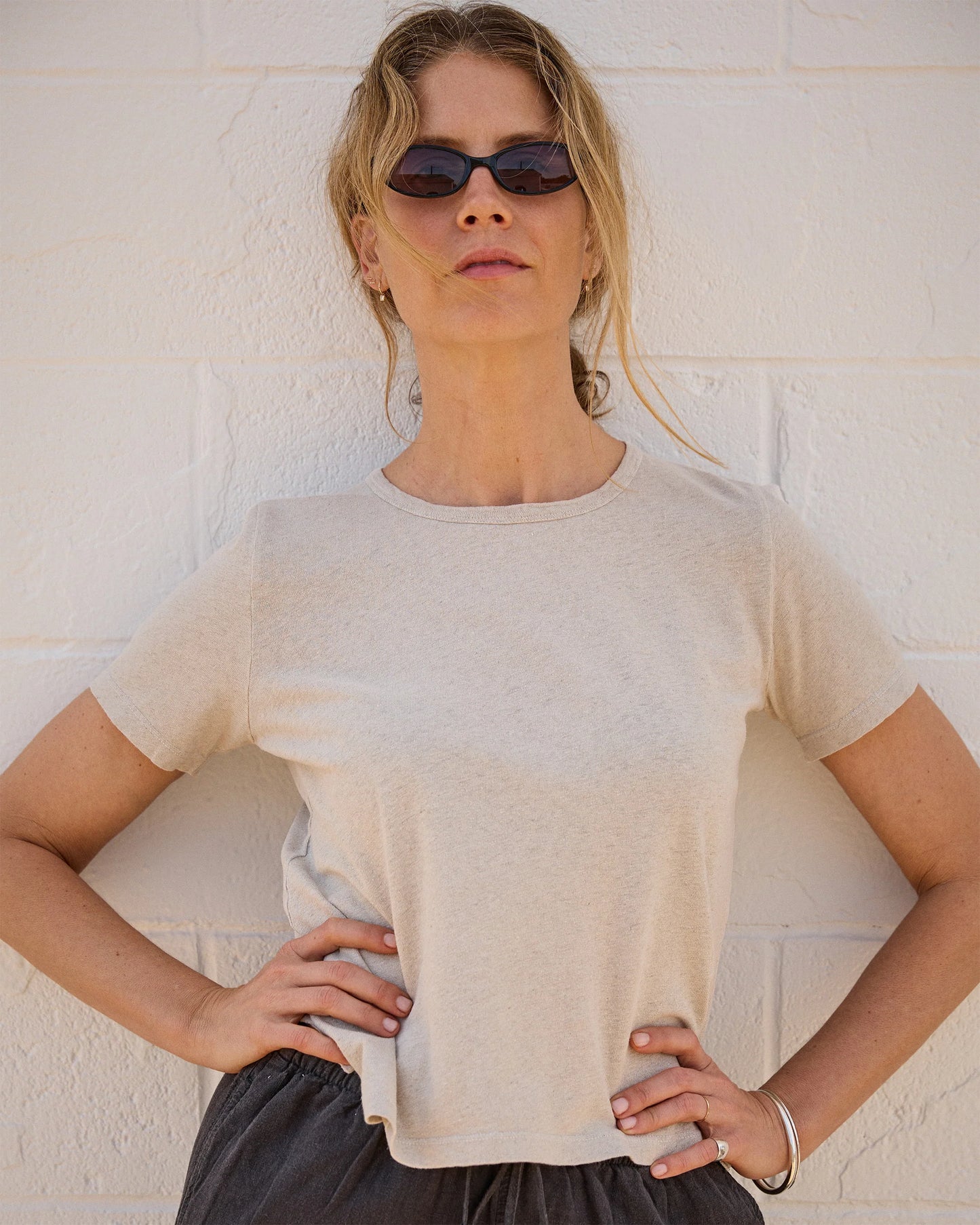 A woman in dark sunglasses, dark pants, and the LinenO Women's Sun Tee in Bone by Alex Crane stands against a white brick wall with her hands on her hips, looking confidently at the camera.